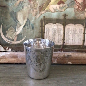 Könnte beinhalten: Eine silberne Tasse mit gravierten floralen und scrollartigen Mustern. Die Tasse hat einen leicht ausgestellten Rand und eine polierte Oberfläche. Der Hintergrund zeigt ein Vintage-Gemälde mit Figuren und einem Kalender.
