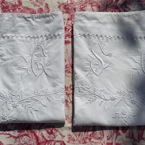 May include: Two white pillowcases with embroidered floral and initial designs. The pillowcases feature an embroidered floral border and the letters "S" and "C". The pillowcases are made of a soft, white fabric.
