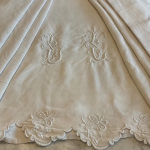 May include: An off-white tablecloth featuring intricate white floral embroidery and monogrammed initials. The fabric has a scalloped edge with detailed needlework patterns along the border and two central embroidered motifs on the draped surface.