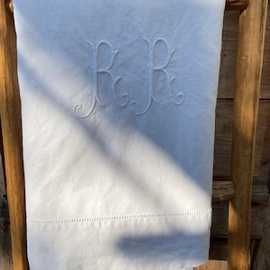 May include: White linen sheet with embroidered initials "RR" in a decorative script. The sheet is draped over a wooden ladder, showcasing the texture and details of the fabric. A row of delicate stitching adorns the bottom edge.