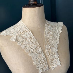 Puede incluir: Un cuello de encaje crema antiguo con un diseño floral. El cuello se exhibe en un maniquí de costura vintage. El encaje presenta patrones detallados de flores y hojas, creando una estética elegante y delicada. El cuello tiene forma de V.