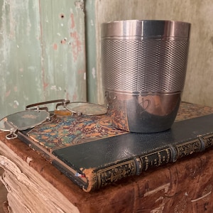 Op de afbeelding: Een zilveren beker met een getextureerd ontwerp en een monogram op de bodem. De beker staat op een groot, antiek boek met een marmeren omslag.