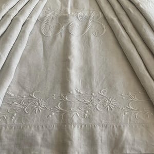 Può includere: Biancheria da letto in lino color crema con dettagli ricamati. La parte superiore presenta un monogramma ricamato, mentre il bordo inferiore mostra un motivo floreale e di vite. Il tessuto è drappeggiato, creando morbide pieghe.