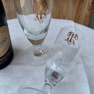May include: Two clear champagne flutes with a gold monogram design. The glasses have a clear stem and base, and a wooden-like accent at the base of the glass. A bottle of Prosecco is partially visible in the background.