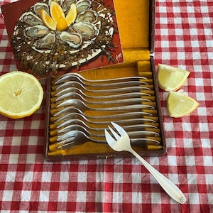 Può includere: Un set di forchette da frutti di mare in metallo argentato disposte in una scatola rettangolare con rivestimento in velluto giallo. Una forchetta è appoggiata fuori dalla scatola. Dietro c'è una carta vintage con ostriche e limoni e spicchi di limone.