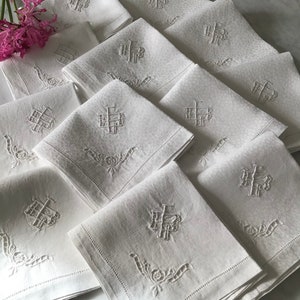 Può includere: Un set di tovaglioli di lino bianco, ciascuno ricamato con un monogramma e un motivo floreale. I tovaglioli sono piegati e impilati, con alcuni visibili in primo piano. Un rametto di fiori rosa si trova nell'angolo in alto a sinistra.