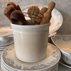 Pode incluir: Um pote cilíndrico de cerâmica na cor creme preenchido com vários utensílios de cozinha de madeira, incluindo pequenas colheres e um cabo, junto com um fino laço de arame escuro, repousando sobre uma pilha de pratos brancos com padrões azuis.