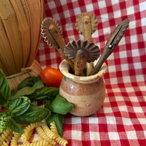 Può includere: Tre rotelle per pasta vintage in legno con bordi metallici seghettati sono inserite in un piccolo vaso di ceramica rotondo color marrone chiaro. Gli utensili da cucina presentano manici in legno usurato e sono disposti verticalmente.