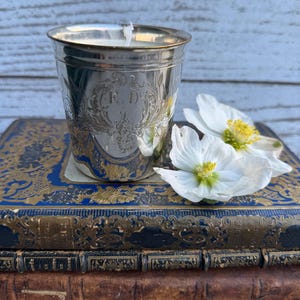 May include: A silver-toned metal cup containing a white candle, featuring an engraved floral motif and the letters E.D. on the front. The cup rests on a stack of two antique books with blue and gold covers, accompanied by two white flowers.