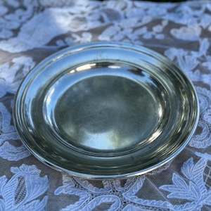 Puede incluir: Un plato redondo de color plateado con un borde ligeramente elevado. El plato tiene una superficie reflectante y está colocado sobre un mantel de encaje blanco. El plato probablemente se utiliza para servir alimentos.