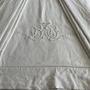 Pode incluir: Tecido de linho creme com iniciais "BB" bordadas em escrita decorativa. O bordado branco apresenta motivos florais e de folhas. O tecido tem uma textura subtil e é drapeado, com uma borda costurada.