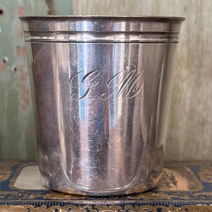 Pode incluir: Uma caneca de metal prateado com um acabamento liso e polido e uma borda ligeiramente alargada. A parte frontal da caneca apresenta as iniciais cursivas gravadas G.M. centradas no corpo. A caneca tem uma forma cilíndrica com base plana.