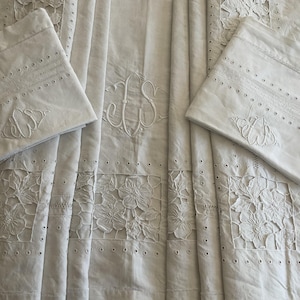 Puede incluir: Juego de cama bordado blanco con dos fundas de almohada y una sábana. La sábana y las fundas de almohada presentan bordados florales y monogramas. Las fundas de almohada tienen costuras decorativas y detalles de ojal. La ropa de cama está hecha de una tela blanca suave.