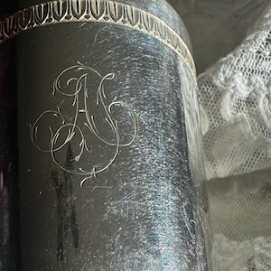 May include: A silver-colored cylindrical container with an ornate, engraved monogram. The container has a decorative band around the top and shows signs of wear. The background is a white, lacy fabric.