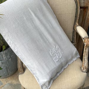 Może przedstawiać: Jasnoszara poszewka na poduszkę z białym haftowanym monogramem, leżąca na krześle w stylu vintage. Poszewka ma falisty brzeg i zapięcie na guzik. Krzesło ma kremowe siedzisko i zdobioną, drewnianą ramę.