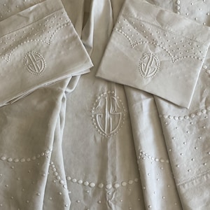 Puede incluir: Juego de ropa de cama de color crema con detalles bordados. El juego incluye dos fundas de almohada y una sábana, todos adornados con un patrón de puntos repetido y un monograma central. Las fundas de almohada tienen un borde festoneado con detalles bordados.