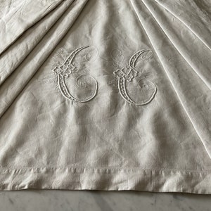 May include: A white linen tablecloth with a delicate embroidered design of two intertwined letters "C" with floral accents.