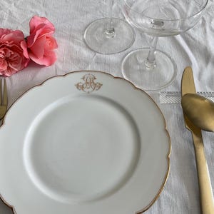 Può includere: Un'elegante mise en place con un piatto bianco con bordo dorato e monogramma. Forchetta, coltello e cucchiaio dorati disposti accanto al piatto. Due calici di champagne e rose rosa completano la composizione.