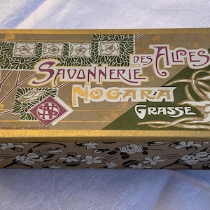 May include: A rectangular cardboard box with a gold-toned lid featuring the text Savonnerie des Alpes Nogara Grasse, decorative green floral patterns, and white edelweiss illustrations. The sides of the box display a repeating black and white floral print.
