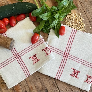 Può includere: Due tovaglioli quadrati in lino color bianco sporco con un motivo a griglia a righe rosse e le lettere monogramma MD ricamate in rosso. I tovaglioli sono presentati con un cetriolo, pomodorini rossi, foglie di basilico fresco e pinoli.