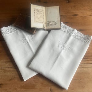 May include: Two white pillowcases with embroidered initials and lace trim are displayed on a wooden surface. An open antique book and a pair of eyeglasses rest on top. The pillowcases are folded, showcasing the delicate embroidery.
