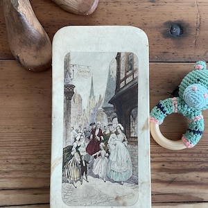 Puede incluir: Una lata rectangular antigua con una ilustración vintage de una escena callejera con figuras. La ilustración presenta tonos apagados y detalles arquitectónicos. También se ven hormas de zapatos de madera y un sonajero de bebé de ganchillo.
