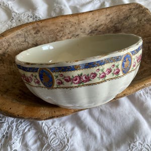 Può includere: Una ciotola in ceramica color crema con bordo smerlato e una fascia decorativa con rose rosa, accenti ovali blu con dettagli dorati e bordo dorato. La ciotola è posizionata all'interno di una madia in legno rustica, allungata e grezza.