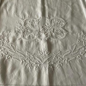 May include: A white textile featuring intricate white embroidery of the letters CB centered above a decorative floral garland. The fabric displays a smooth texture with subtle folds and a stitched hem along the bottom edge of the piece.