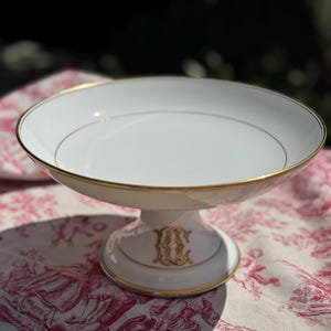 Op de afbeelding: Een witte porseleinen taartstandaard met een gouden rand en een gouden monogram. De standaard staat op een rood-wit patroon tafelkleed. De taartstandaard heeft een diameter van ongeveer 20 cm.