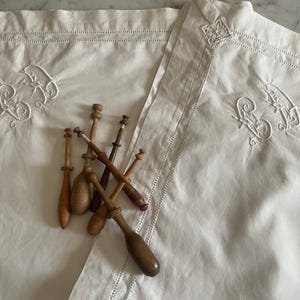 Puede incluir: Fundas de almohada de lino blanco con iniciales bordadas y costuras decorativas. Una colección de bolillos de madera descansa sobre las fundas de almohada. Los bolillos varían en tamaño y tono de marrón, sugiriendo un aspecto vintage.