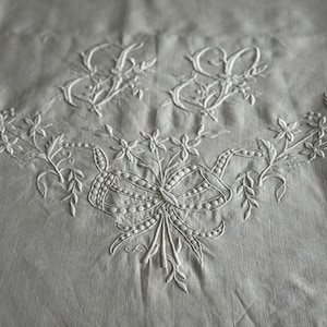 Puede incluir: Primer plano de un textil gris claro con bordados blancos de diseños florales y de cintas. Dos iniciales ornamentadas en la parte superior, con una guirnalda de flores y hojas debajo. El bordado es detallado y delicado.