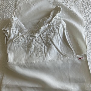 May include: White vintage-style nightgown and pillowcase set. The nightgown features floral embroidery on the bodice and delicate lace trim. The pillowcase has a scalloped edge and matching embroidery. The set is made of soft, white fabric.