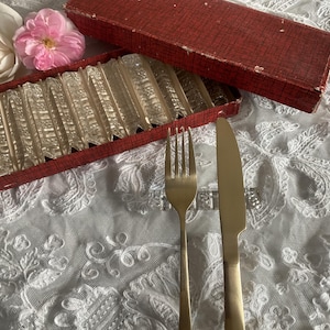 Può includere: Un set di coltelli vintage in vetro trasparente in una scatola rossa, con una forchetta e un coltello dorati su una tovaglia di pizzo bianco. Due rose, una bianca e una rosa, sono sullo sfondo. I coltelli sono in una scatola rossa.