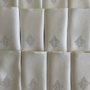 Könnte beinhalten: Eine Sammlung gefalteter weißer Leinen-Servietten. Jede Serviette hat ein gesticktes, verziertes rautenförmiges Design in einem helleren Weißton. Die Servietten sind ordentlich angeordnet und zeigen ihre elegante Textur und ihr Design.