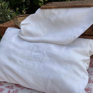 May include: Two white rectangular pillowcases made of fabric, each featuring an embroidered monogram with the letters BG in a cursive script. The pillowcases have small white buttons along the side edge for closure and a smooth, plain texture.