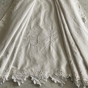 Puede incluir: Textil de lino blanco con iniciales bordadas "UP" y un borde floral festoneado. La tela está drapeada, mostrando la textura y los detalles del bordado. La estética general es vintage y elegante.