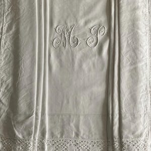 Può includere: Lenzuolo bianco in lino con le iniziali ricamate "M P" in una scrittura decorativa. Il lenzuolo presenta un delicato bordo in pizzo e pieghe verticali. Il tessuto ha una trama sottile, che suggerisce un pezzo vintage o antico.