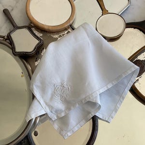 以下が含まれることがあります： 刺繍された花柄の白いリネンハンカチが、様々なフレームスタイルと形状のアンティーク手鏡に囲まれた銀色のトレイの上に置かれています。鏡は周囲の物体を反射しています。