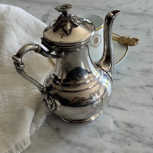 Puede incluir: Una tetera de plata pulida con asa curva, pico alto y tapa rematada con un adorno floral. El cuerpo de la tetera presenta un monograma grabado. Detrás se observa una taza de porcelana blanca con borde dorado y un monograma en color oro.