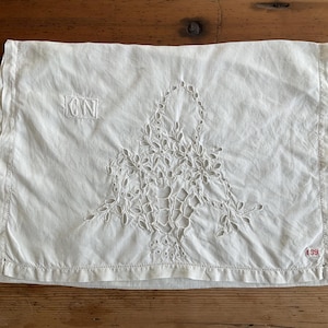 Puede incluir: Funda de almohada rectangular de color crema con intrincado diseño floral calado. Presenta las iniciales "CN" en un cuadrado en la parte superior y el número "39" en rojo en la parte inferior. La funda está hecha de una tela suave y ligeramente arrugada.