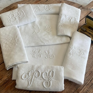 May include: A selection of white linen textiles, including folded napkins and a larger sheet, all embroidered with elegant, ornate monograms. The embroidery is in a matching white thread, creating a subtle, textured effect. The items are arranged on a rustic wooden surface.