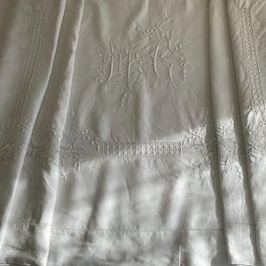 Op de afbeelding: Wit linnen laken met geborduurde bloemen en initialen. Het laken heeft delicate bloempatronen en de initialen "MGC" in het midden, met decoratieve randen langs de randen. De stof is gevouwen, wat de textuur en het ontwerp laat zien.