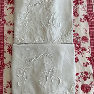 May include: Two white rectangular pillowcases featuring white embroidered details. Each case displays the text "BUONI RIPOSO" in a curved banner above the ornate, intertwined letters "F" and "A," with a floral vine pattern along the bottom edge.