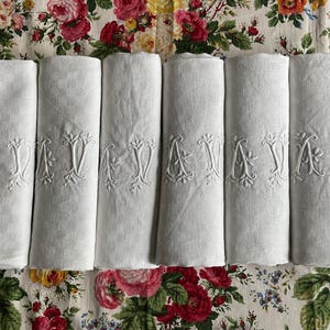 Op de afbeelding: Zes opgerolde witte linnen servetten met geborduurde "AV" monogrammen. De servetten liggen op een bloemmotiefstof met rode, roze en gele rozen. De servetten zijn een klassieke toevoeging aan elke tafel.