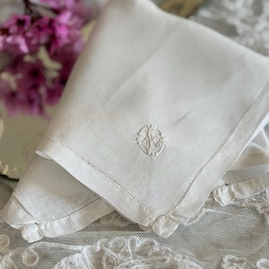 Puede incluir: Un pañuelo de lino blanco doblado con un monograma bordado en la esquina. Los bordes están acabados con un delicado pespunte y pequeños volantes. Un espejo y flores rosas están en el fondo.