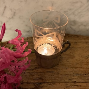 May include: A lit candle inside a clear glass votive holder with etched bow and floral designs. The holder sits in a silver-toned metal cup with a handle. Pink flowers are in the foreground, and the background is a wooden surface.