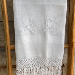 Könnte beinhalten: Weißes Leinenhandtuch mit gestickten Initialen und dekorativer Fransen. Das Handtuch ist über eine Holzleiter drapiert. Die Initialen sind in zarter Schrift. Die Fransen sind ein gewebtes Design.