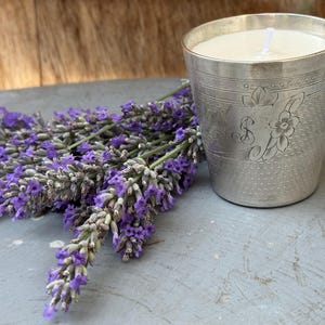 May include: A silver candle holder with a white candle and a bundle of purple lavender. The candle holder has an engraved floral design. The lavender is a vibrant purple color.