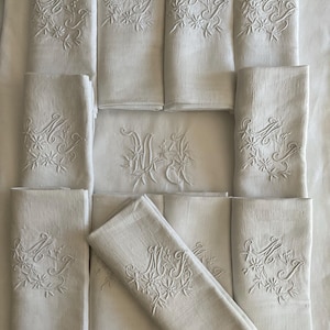 Pode incluir: Uma coleção de guardanapos de tecido branco retangulares, cada um com um monograma bordado das letras MJ rodeado por motivos florais. Os guardanapos estão dispostos numa grelha, com um dobrado diagonalmente no centro.