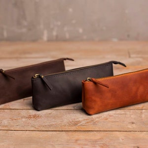 May include: Three leather pencil cases in brown, black, and tan. The cases are lined up on a wooden surface.